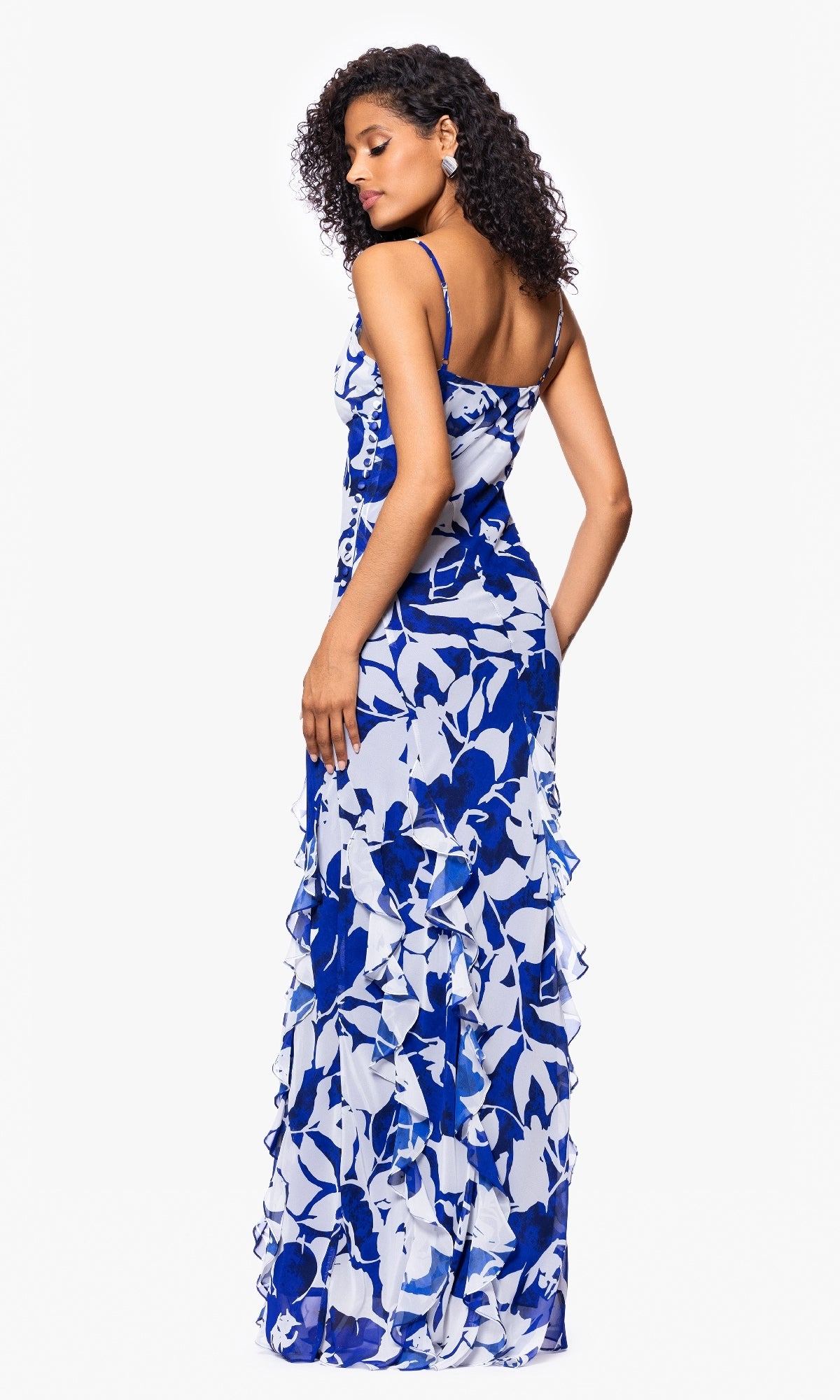 A woman models the Betsy and Adam A26915 Long Formal Dress, a blue and white floral sleeveless maxi with ruffle detailing, as she poses with her back slightly turned against a plain white background.